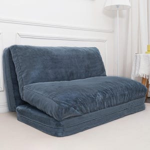 Blue plush foldable sofa or futon with a padded backrest and seat in a cozy room setting, featuring a lamp and side table.