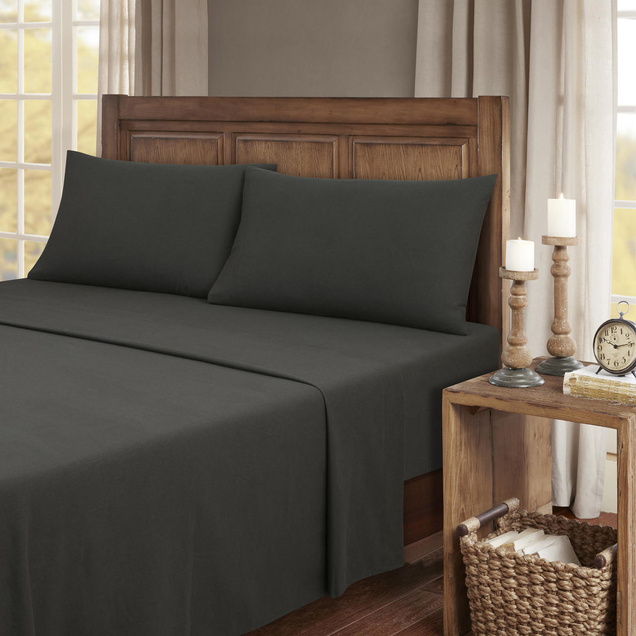 Dark gray bed sheets and pillowcases on a wooden bed, with a wooden nightstand featuring candles, a clock, and books in a woven basket.