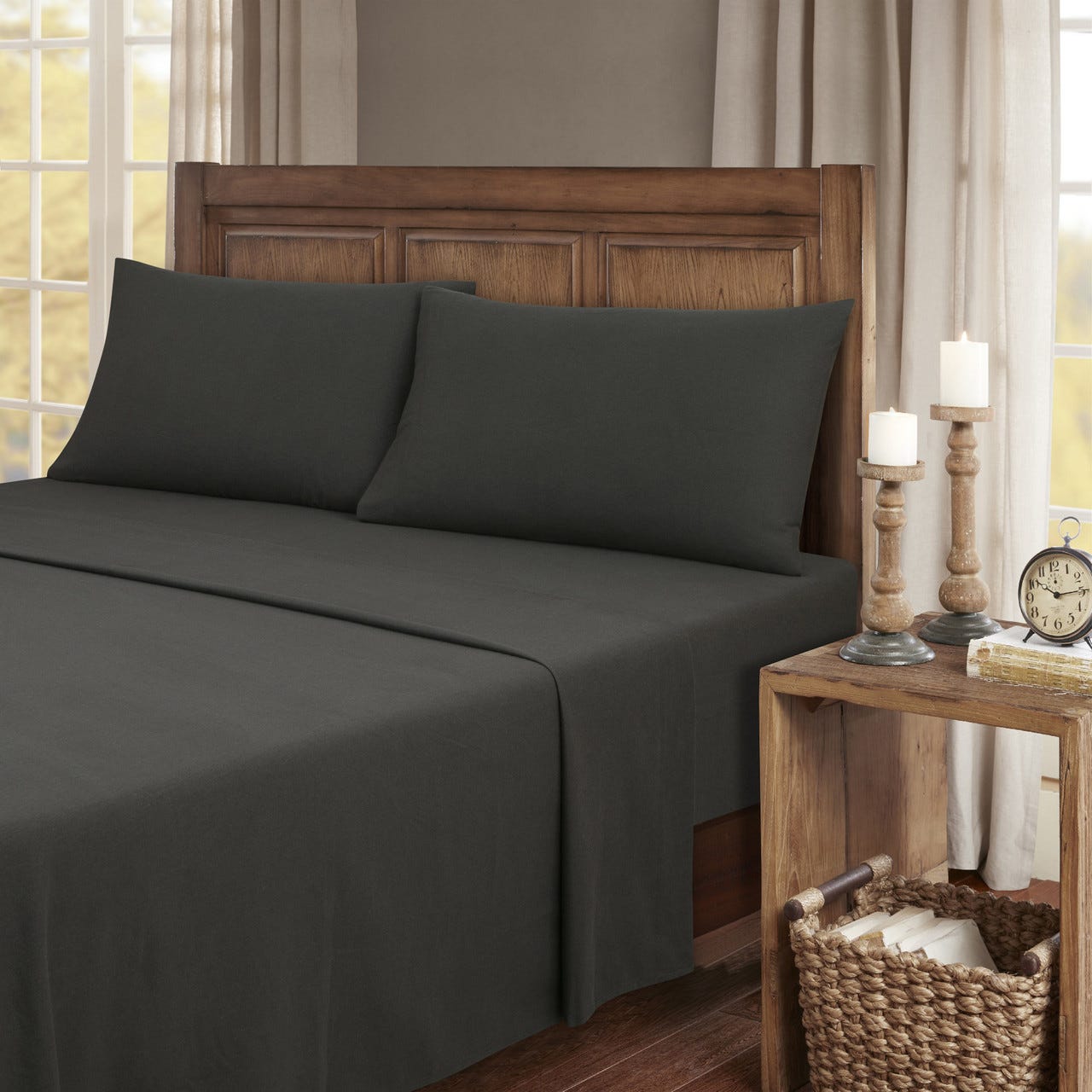 Dark gray bed sheets and pillowcases on a wooden bed, with a wooden nightstand featuring candles, a clock, and books in a woven basket.