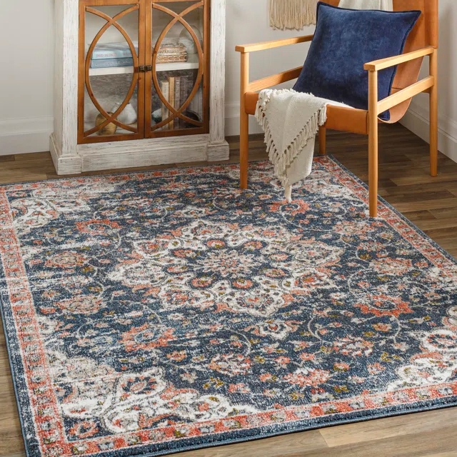 The Bungalow Rose Floral Area Rug features a traditional floral pattern with a mix of blue, orange, and white colors. It rests on a hardwood floor next to a wooden chair with a blue cushion and a textured blanket draped over the arm.