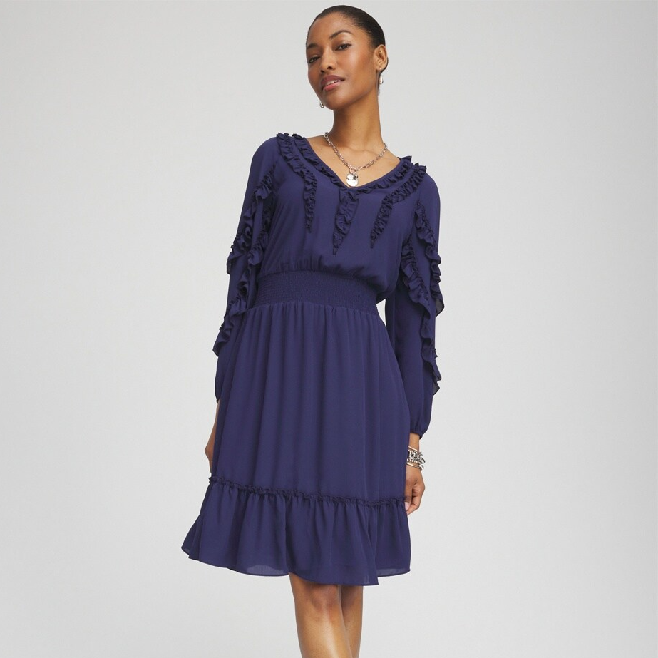 A woman is wearing a navy blue dress with ruffled details and long sleeves, accessorized with a necklace and earrings.