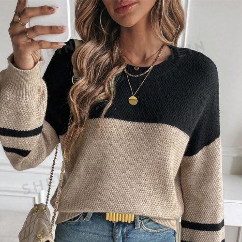 A woman is wearing a striped color-block sweater with a gold pendant necklace, holding a smartphone, and sporting a cream handbag and a yellow belt.