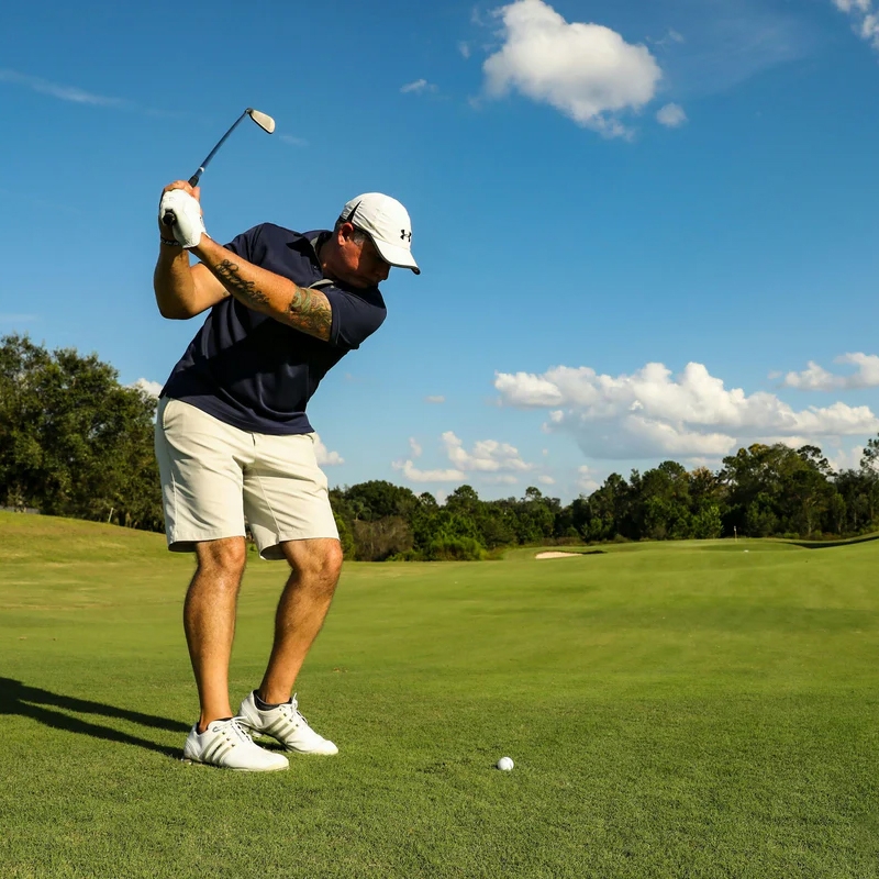 A golfer in white shorts, a navy shirt, and a white Under Armour cap is preparing to swing. He is wearing white Adidas golf shoes on a lush green fairway under a bright blue sky.