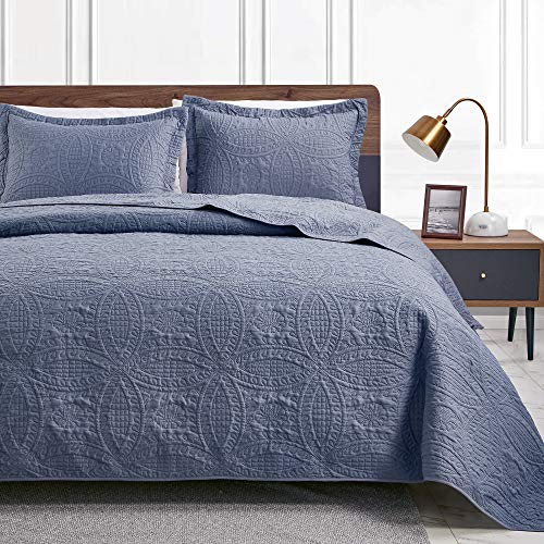 A blue quilt with detailed pattern stitching is spread on a bed with two matching pillow shams set against a wooden headboard and a nightstand with a lamp.
