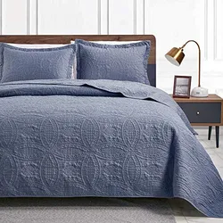 A blue quilt with detailed pattern stitching is spread on a bed with two matching pillow shams set against a wooden headboard and a nightstand with a lamp.