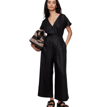 A woman is wearing a black jumpsuit with a V-neck and wide legs, paired with black sandals. She's holding a textured handbag.