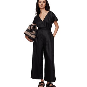 A woman is wearing a black jumpsuit with a V-neck and wide legs, paired with black sandals. She's holding a textured handbag.