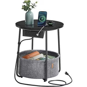 Round side table with a black top, featuring built-in power outlets and USB ports. It includes a gray fabric storage basket underneath and holds a vase, phone, and watch on top.