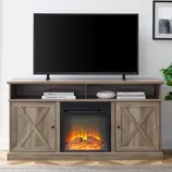 A brown wooden TV stand with farmhouse-style doors features an electric fireplace insert, a flat-screen TV on top, and shelves with books and decor items.