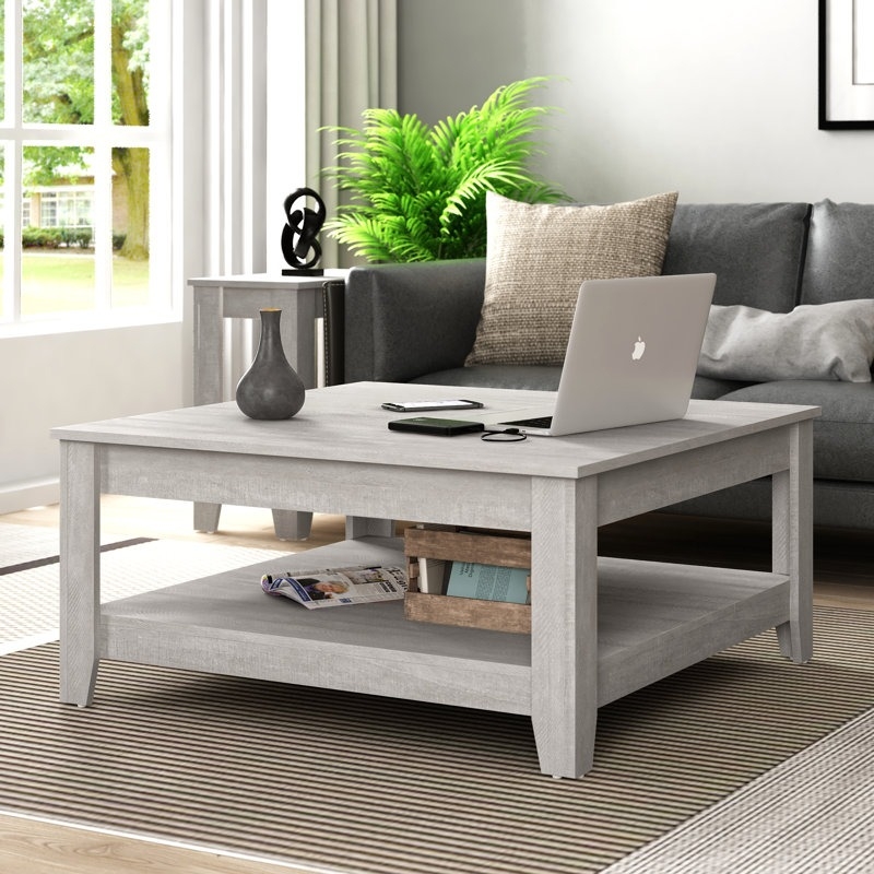 A gray wooden coffee table with a lower shelf holds a laptop, phone, and vase in a cozy living room. It is complemented by a gray sofa, a plant, and a side table with a decorative sculpture.