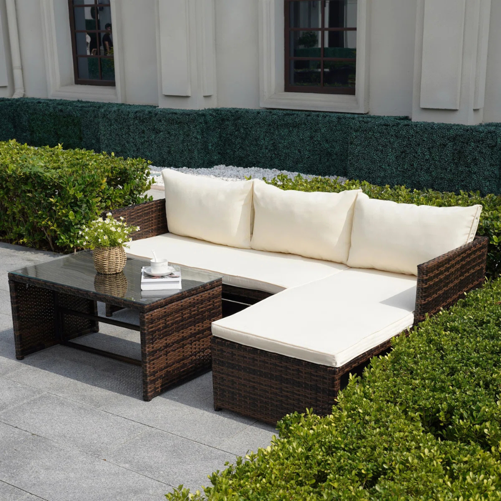 Outdoor patio set with an L-shaped cushioned sofa in beige and a glass-top coffee table, both made from dark brown wicker.