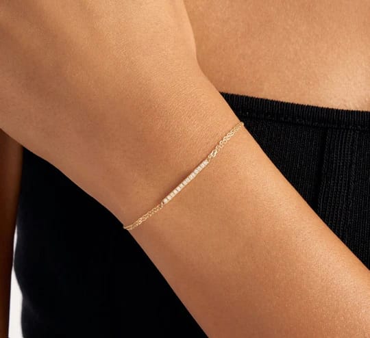 A delicate gold chain bracelet with a small row of tiny white beads worn on a person's wrist, paired with a black strapless top.