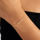 A delicate gold chain bracelet with a small row of tiny white beads worn on a person's wrist, paired with a black strapless top.