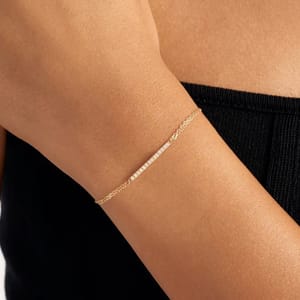 A delicate gold chain bracelet with a small row of tiny white beads worn on a person's wrist, paired with a black strapless top.
