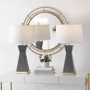 Two modern table lamps with dark gray bases and white shades are placed on a white console table, flanking a small vase of flowers and books. A round, gold-framed mirror is mounted on the wall above.