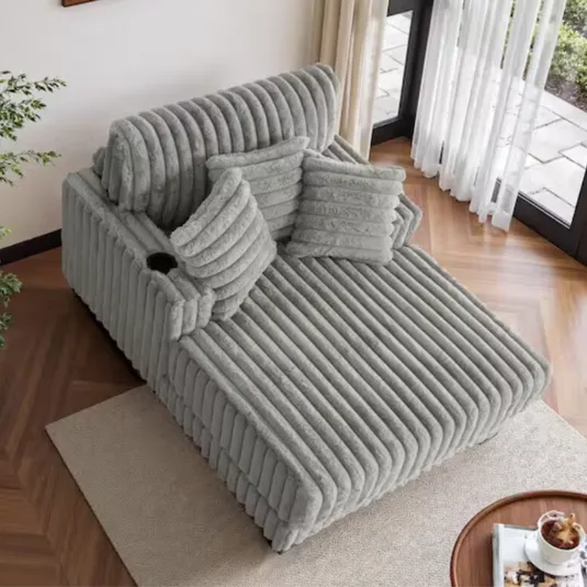 Gray, ribbed chaise lounge with matching throw pillows, cup holder, and positioned on a neutral rug by a window.