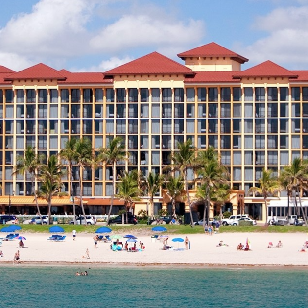 A beachfront hotel with a red roof stands behind a sandy beach dotted with blue umbrellas and palm trees. People are enjoying the sun and ocean.