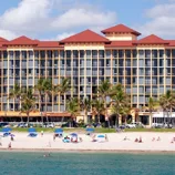 A beachfront hotel with a red roof stands behind a sandy beach dotted with blue umbrellas and palm trees. People are enjoying the sun and ocean.