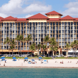 A beachfront hotel with a red roof stands behind a sandy beach dotted with blue umbrellas and palm trees. People are enjoying the sun and ocean.