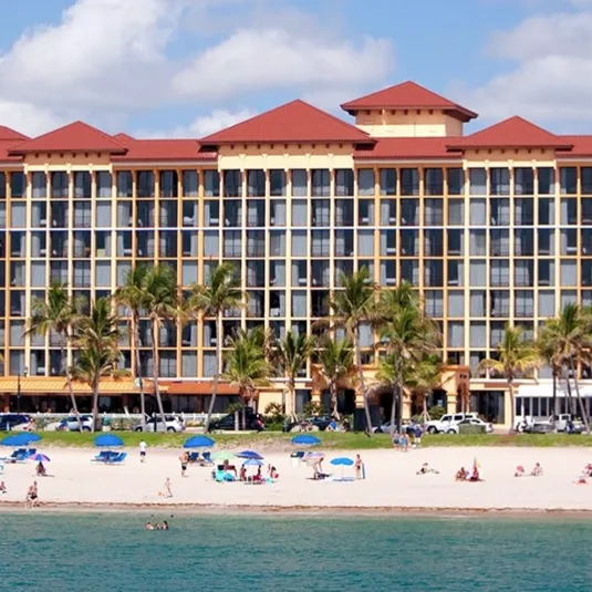 A beachfront hotel with a red roof stands behind a sandy beach dotted with blue umbrellas and palm trees. People are enjoying the sun and ocean.