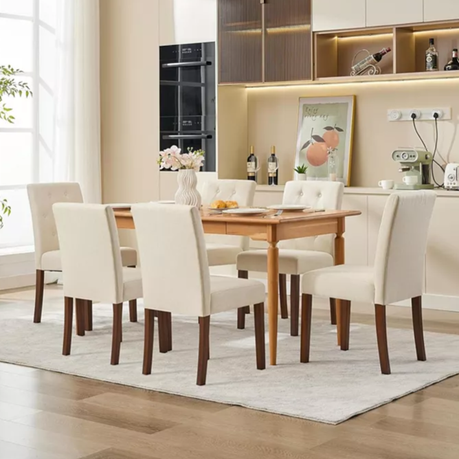 A dining room set with a wooden table and six upholstered chairs in a neutral cream color. The background features a modern kitchen with light cabinetry, built-in appliances, and decorative elements like a vase, framed artwork, and wine bottles.