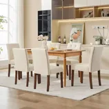 A dining room set with a wooden table and six upholstered chairs in a neutral cream color. The background features a modern kitchen with light cabinetry, built-in appliances, and decorative elements like a vase, framed artwork, and wine bottles.