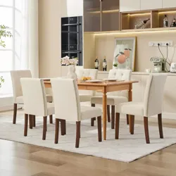 A dining room set with a wooden table and six upholstered chairs in a neutral cream color. The background features a modern kitchen with light cabinetry, built-in appliances, and decorative elements like a vase, framed artwork, and wine bottles.