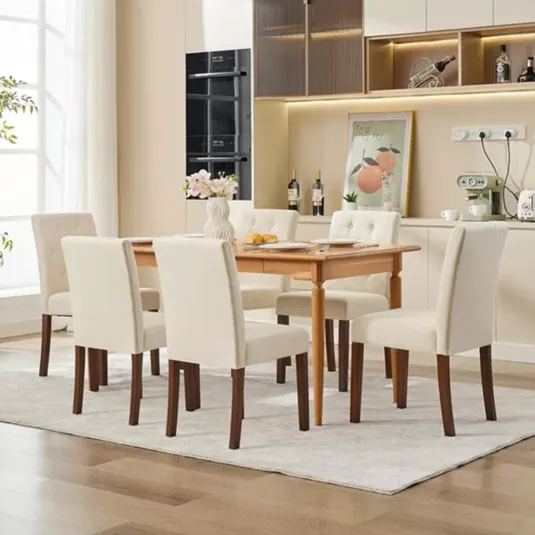 A dining room set with a wooden table and six upholstered chairs in a neutral cream color. The background features a modern kitchen with light cabinetry, built-in appliances, and decorative elements like a vase, framed artwork, and wine bottles.