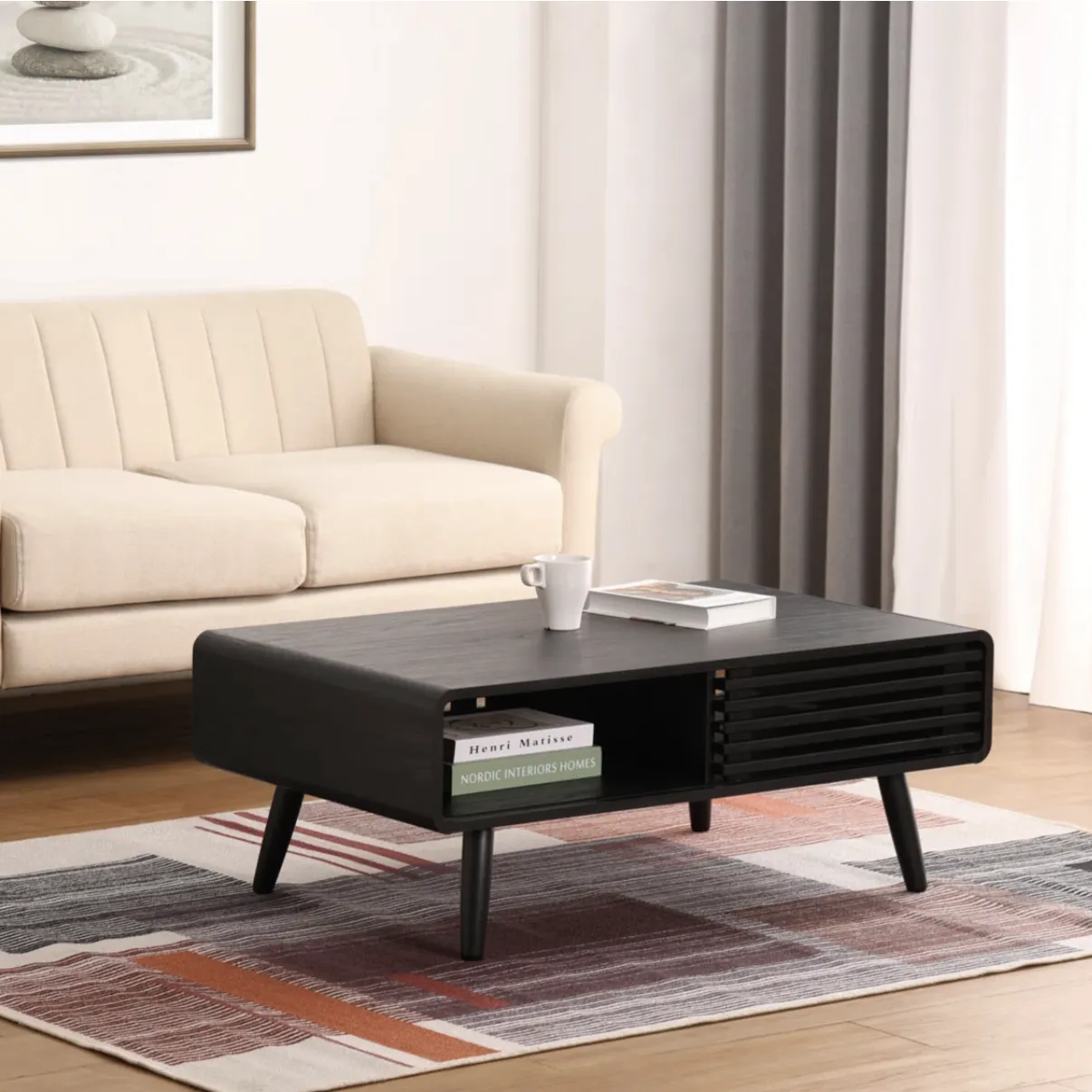 A modern living room features a beige sofa, a black rectangular coffee table with storage, and a patterned area rug. The coffee table holds books and a white mug, while the wall displays a framed zen-like artwork.