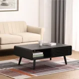 A modern living room features a beige sofa, a black rectangular coffee table with storage, and a patterned area rug. The coffee table holds books and a white mug, while the wall displays a framed zen-like artwork.
