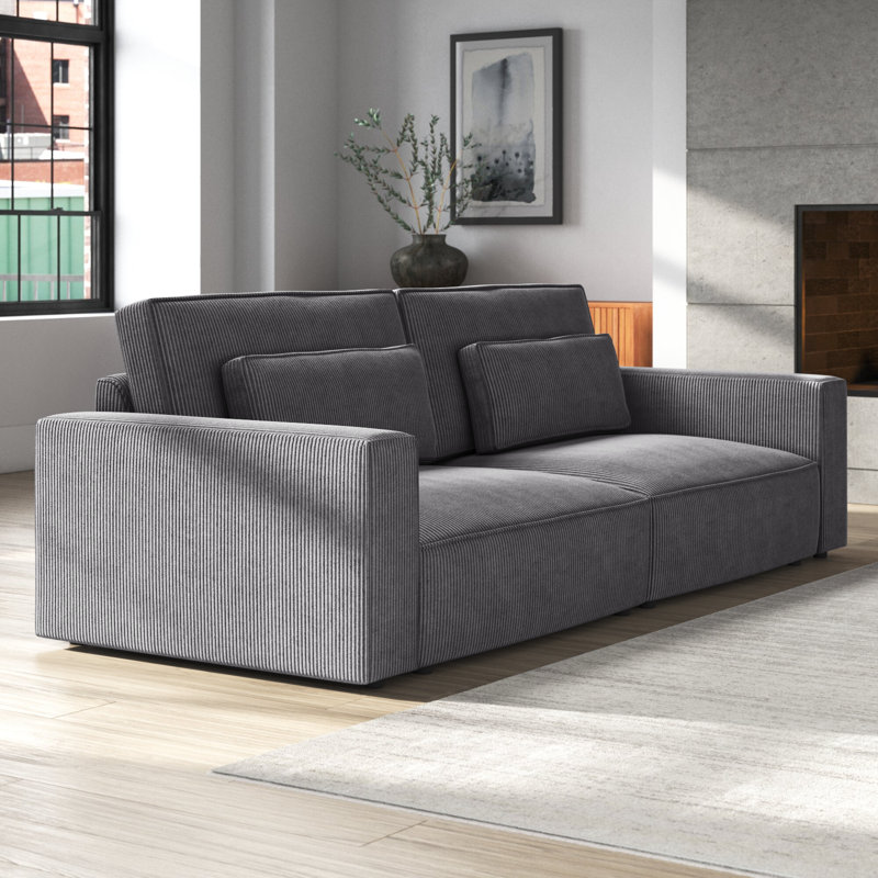 A gray corduroy sofa with an extra-deep design, featuring wide armrests and layered cushions for added comfort. The sofa is set in a modern living room with a neutral color palette and large windows.
