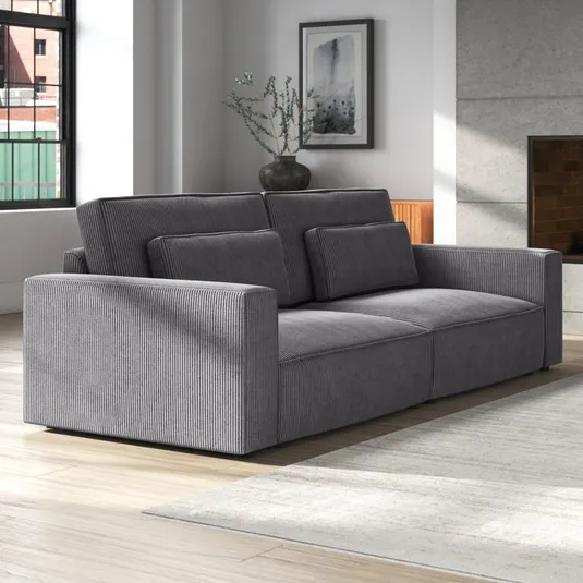 A gray corduroy sofa with an extra-deep design, featuring wide armrests and layered cushions for added comfort. The sofa is set in a modern living room with a neutral color palette and large windows.