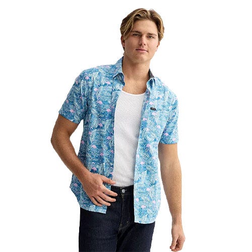 A man wears a blue short-sleeve button-up shirt with a tropical print over a white tank top and dark jeans.