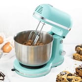 A teal stand mixer with a stainless-steel bowl and whisk attachment, surrounded by eggs and cookie ingredients.