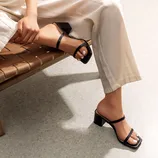 A person is sitting on a woven bench wearing black strappy heeled sandals and white tailored pants.