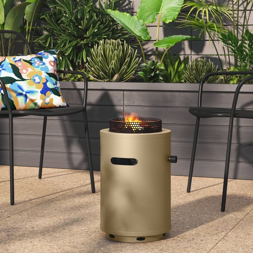A small outdoor fire pit is on a patio surrounded by two black chairs, one with a colorful floral cushion. Lush green plants are in the background.