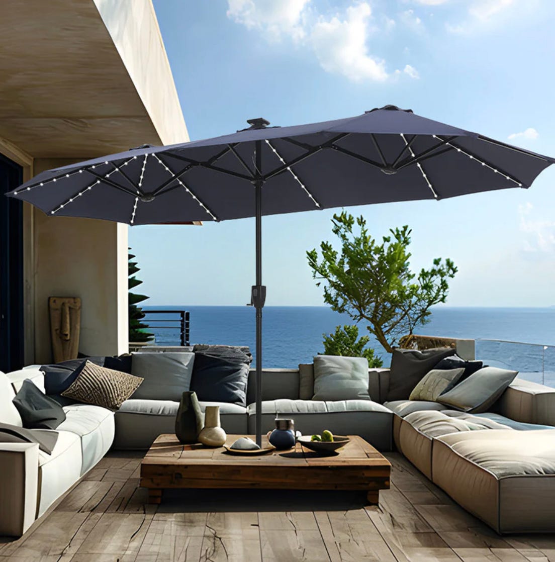 Large outdoor umbrella with a cantilever design, surrounded by modular patio furniture including sofas and a wooden coffee table, overlooking the sea.
