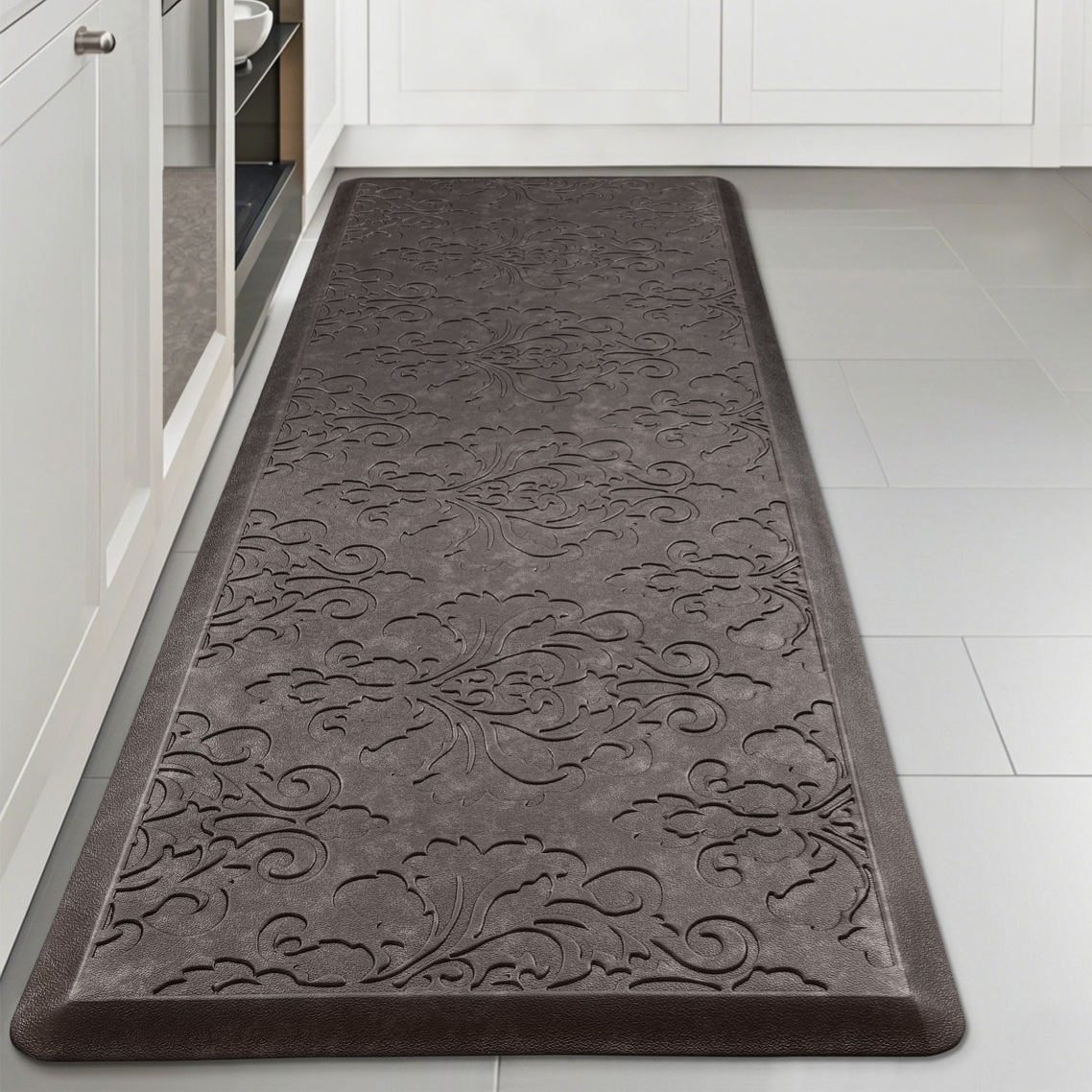 A gray anti-fatigue mat with an embossed floral pattern on a kitchen floor, placed between white cabinets and an appliance.
