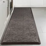 A gray anti-fatigue mat with an embossed floral pattern on a kitchen floor, placed between white cabinets and an appliance.