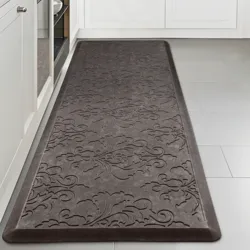 A gray anti-fatigue mat with an embossed floral pattern on a kitchen floor, placed between white cabinets and an appliance.