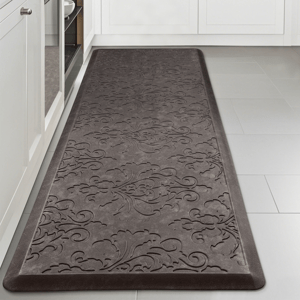 A gray anti-fatigue mat with an embossed floral pattern on a kitchen floor, placed between white cabinets and an appliance.