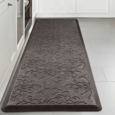 A gray anti-fatigue mat with an embossed floral pattern on a kitchen floor, placed between white cabinets and an appliance.