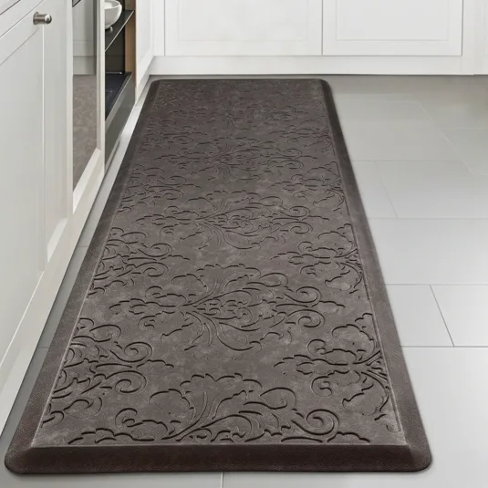 A gray anti-fatigue mat with an embossed floral pattern on a kitchen floor, placed between white cabinets and an appliance.