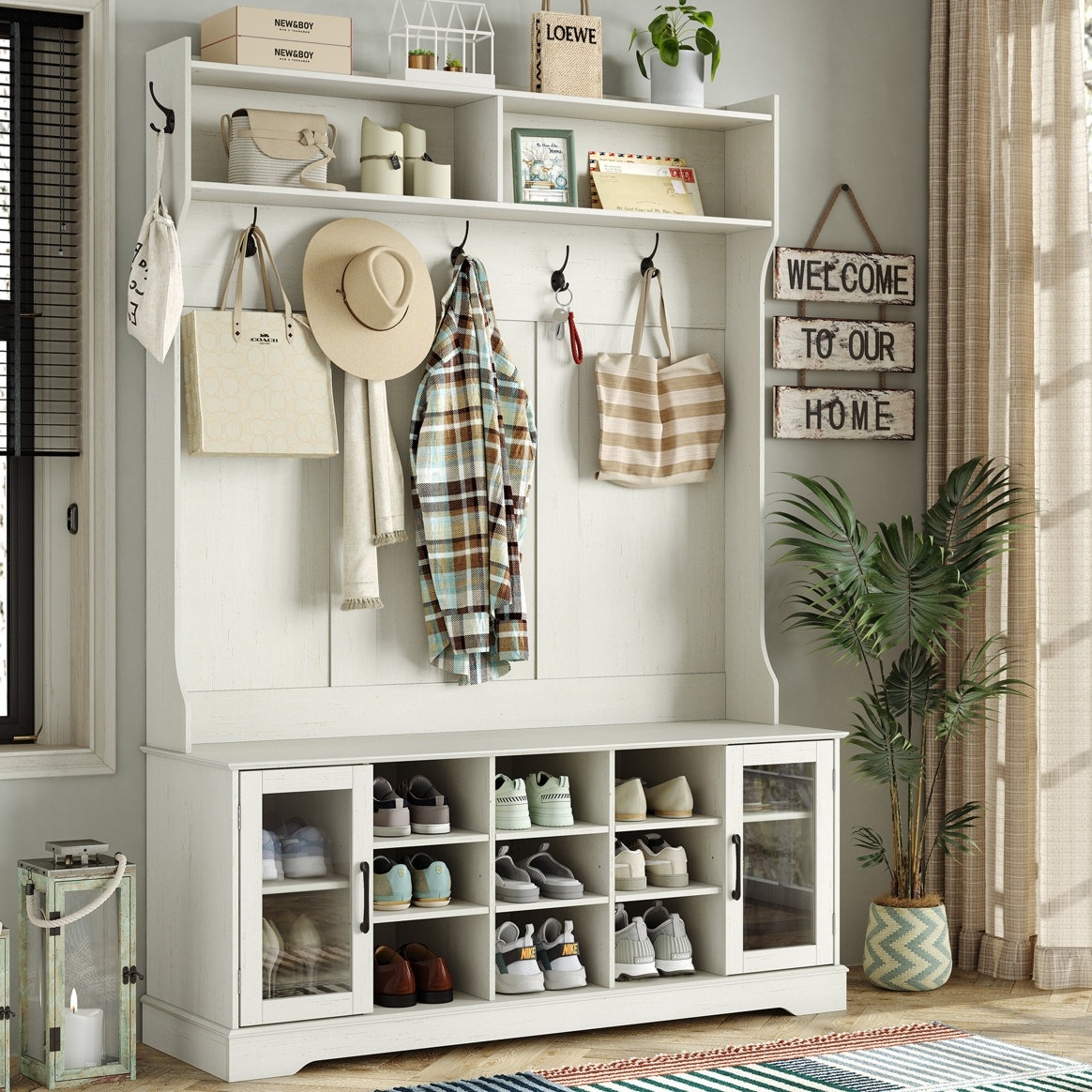 Entryway storage unit with shelves, hooks, and cubbies for shoes. Features hats, bags, a plaid shirt, and decor items like candles and plants. A sign reads \“Welcome to Our Home,\“ and there's a lantern and potted plant nearby.