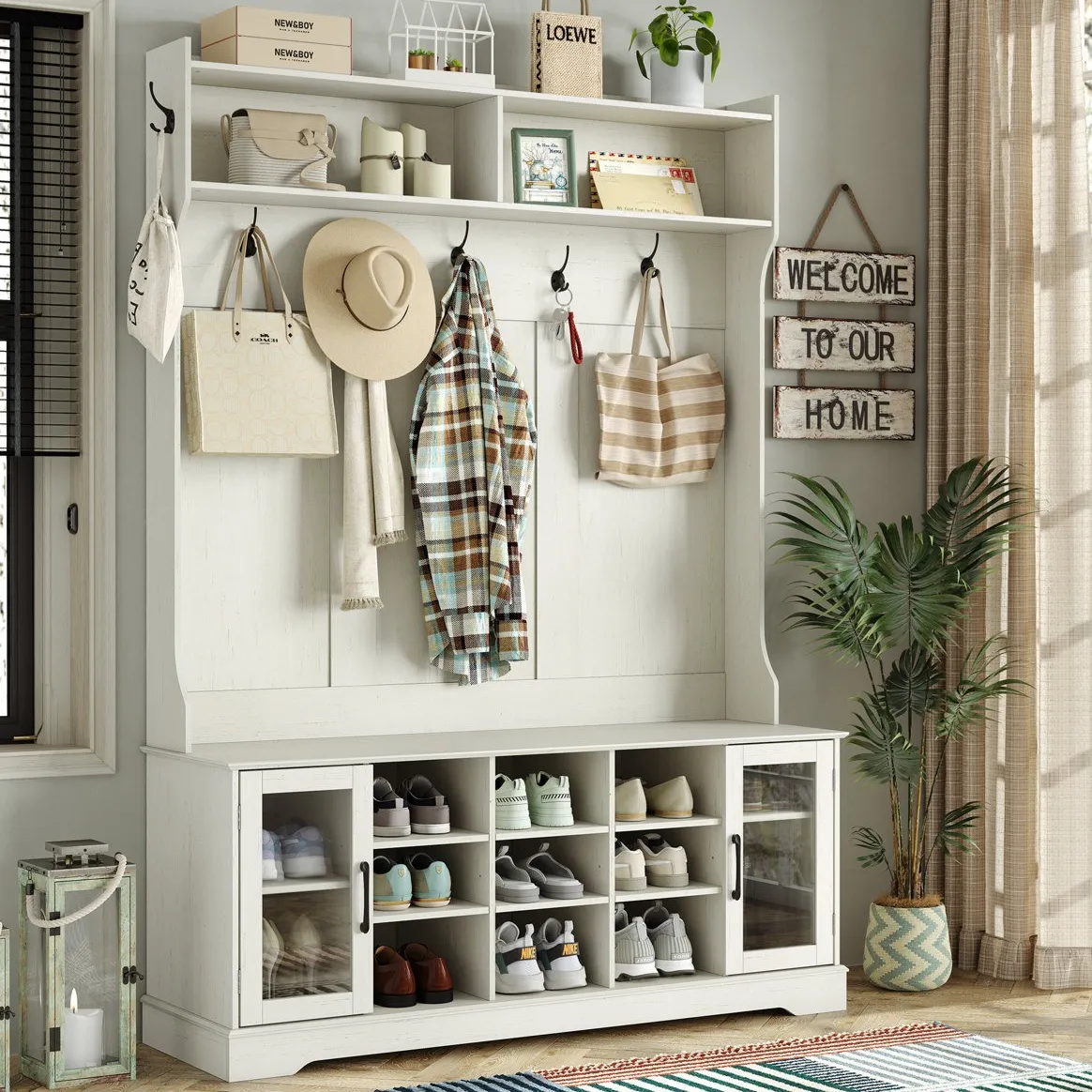 Entryway storage unit with shelves, hooks, and cubbies for shoes. Features hats, bags, a plaid shirt, and decor items like candles and plants. A sign reads \“Welcome to Our Home,\“ and there's a lantern and potted plant nearby.