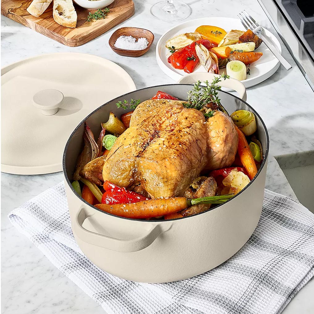 A beige Dutch oven containing a roasted chicken and vegetables sits on a white checkered cloth. Nearby, there's a wooden board with bread and a plate with cooked vegetables.