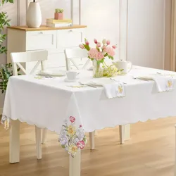 A dining table is set with floral embroidered tablecloth and matching napkins, featuring elegant place settings. A vase with pink flowers serves as a centerpiece, while a cabinet with books and a lamp is visible in the background.