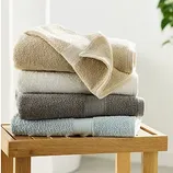 A stack of neatly folded towels in beige, white, gray, and light blue is placed on a wooden bench, with a small plant partially visible in the background.