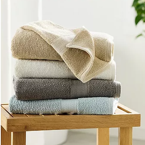 A stack of neatly folded towels in beige, white, gray, and light blue is placed on a wooden bench, with a small plant partially visible in the background.