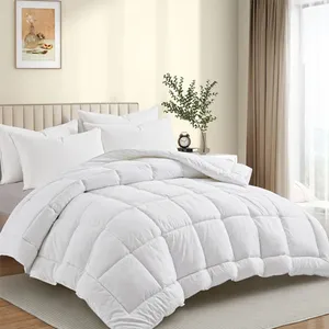 A white, quilted all-season down alternative comforter laid on a neatly made bed with a beige headboard, surrounded by white pillows. The bedroom features a light color palette with a framed art piece and a vase of greenery in the background.